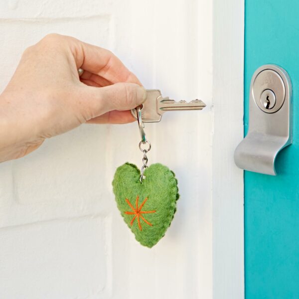 A hand is holding a key with a green, Felt Heart Keyring on its keyring, about to unlock a door. The key is inserted into a silver lock on a teal-colored door.