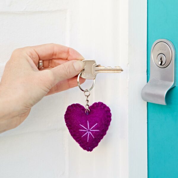 A hand holding a key with a Felt Heart Keyring is inserting the key into a silver lock on a bright turquoise door.