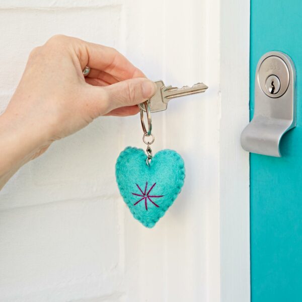 A hand holding a key with a Felt Heart Keyring dangles from it, about to insert it into a door lock near a turquoise door frame.