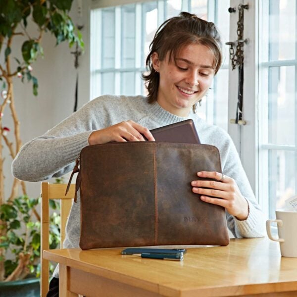 Someone at a table places a tablet into a Personalised Buffalo Leather 14 inch Laptop Case, with a smartphone, pen, and mug nearby.