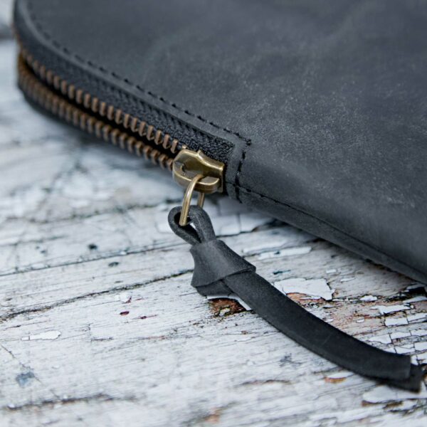 Close-up of the Personalised Buffalo Leather 14 inch Laptop Case in black with a brass zipper and leather pull tab, shown on a weathered white wooden surface—perfect for protecting your laptop in style.