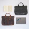 Two Personalised Buffalo Leather 15 inch Laptop Cases, one in dark brown and one in black, are displayed on a light surface next to an open book, a closed laptop, and a pair of glasses.