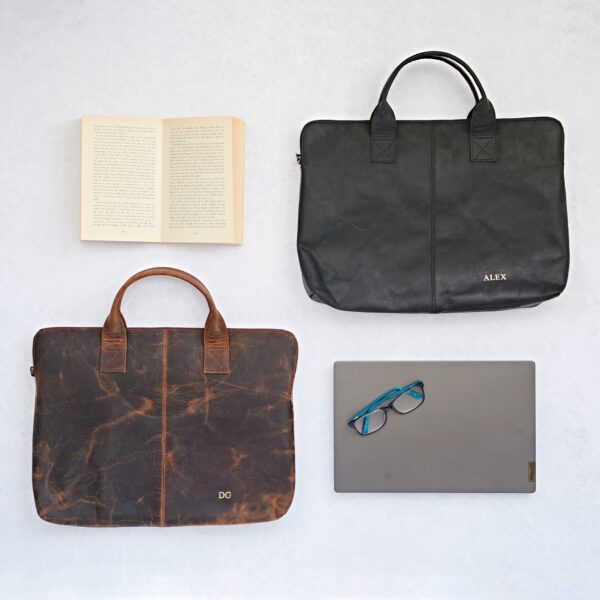 Two Personalised Buffalo Leather 15 inch Laptop Cases, one in dark brown and one in black, are displayed on a light surface next to an open book, a closed laptop, and a pair of glasses.