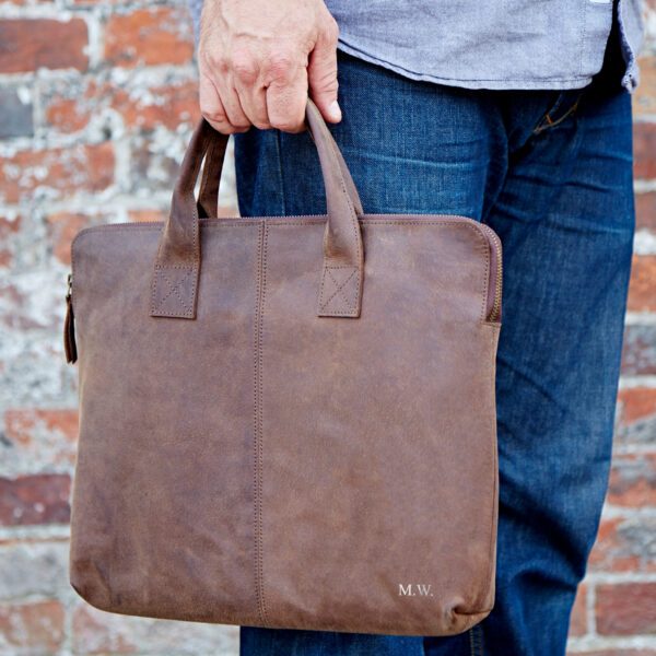 A person wearing jeans stands before a brick wall, holding a Personalised Buffalo Leather 15 inch Laptop Case with two handles and "M.W." initials embossed on the lower corner.