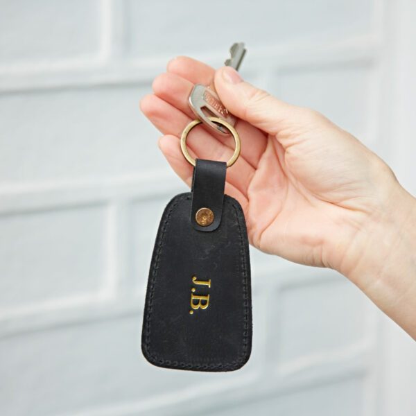 A hand holds a key attached to a black buffalo leather tag keyring with gold initials "J.B." embossed on it.