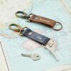 Two Personalised Buffalo Leather Keyrings with Hook, labeled "MADDIE" and "TOM," hold keys atop a world map—elegant, quality leather mementos of adventure.