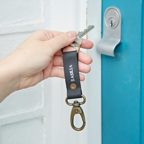 A hand holds a key with a Black Leather Keyring labelled "SASKIA" near a blue door and silver lock.