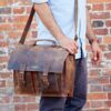 A person in a light blue shirt carries the Personalised Brown Buffalo Leather Satchel Style Briefcase with buckles and a shoulder strap, standing before a brick wall.