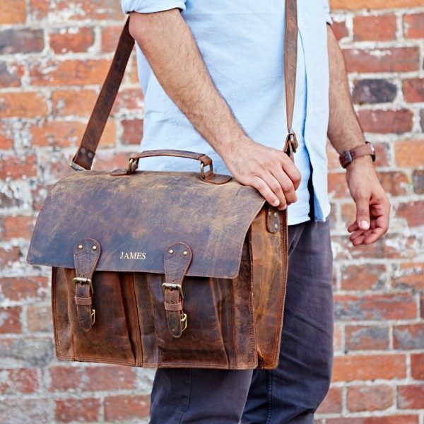 A person in a light blue shirt carries the Personalised Brown Buffalo Leather Satchel Style Briefcase with buckles and a shoulder strap, standing before a brick wall.