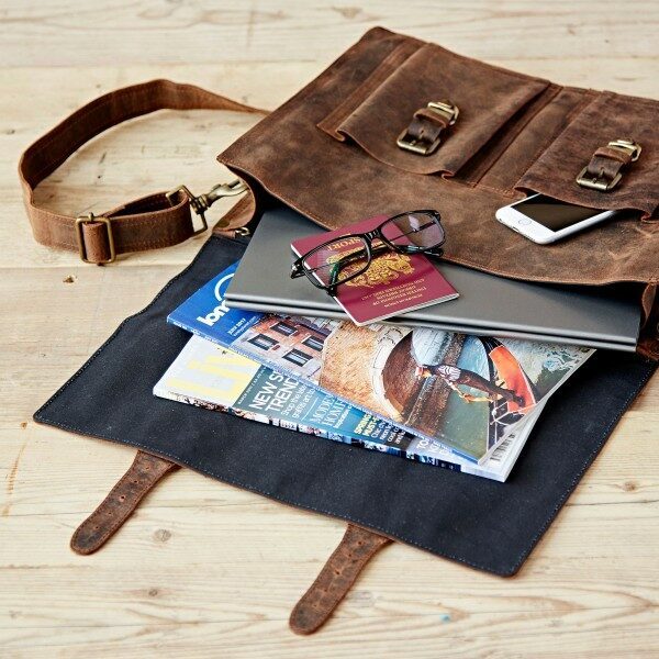 A Personalised Brown Buffalo Leather Satchel Style Briefcase sits on a wooden surface, partially open to reveal a laptop, phone, glasses, passport, brochure, and magazines inside.