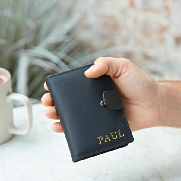 A hand holds the Personalised Buffalo Leather Credit Card Wallet in black, featuring a snap closure and "PAUL" embossed in gold, placed beside a white mug on a light surface.