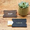 Two Personalised Buffalo Leather Travel Card Holders—one brown embossed "DAN" and one black with "L.K.D"—rest on a wooden surface next to a small potted succulent and a train ticket.