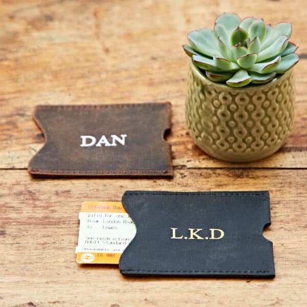 Two Personalised Buffalo Leather Travel Card Holders—one brown embossed "DAN" and one black with "L.K.D"—rest on a wooden surface next to a small potted succulent and a train ticket.