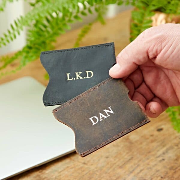 A hand holds two Personalised Buffalo Leather Travel Card Holders—one black with "L.K.D" and one brown with "DAN" embossed on the front. A laptop and a plant are visible in the background.
