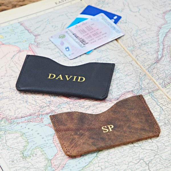 Two Personalised Buffalo Leather Slim Card Holders—one black with "DAVID" and one brown with "SP"—are displayed on a map alongside ID and credit cards.