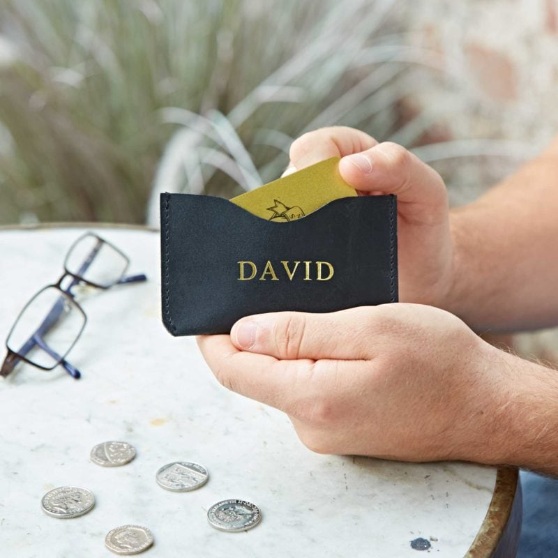 Someone inserts a card into the Personalised Buffalo Leather Slim Card Holder with "DAVID" printed on it, while coins and eyeglasses sit on a round white table.