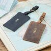 Two Personalised Buffalo Leather Luggage Tags, one black monogrammed “NK” and one brown with “WJA,” are displayed on an open atlas, with a passport partly visible in the background.