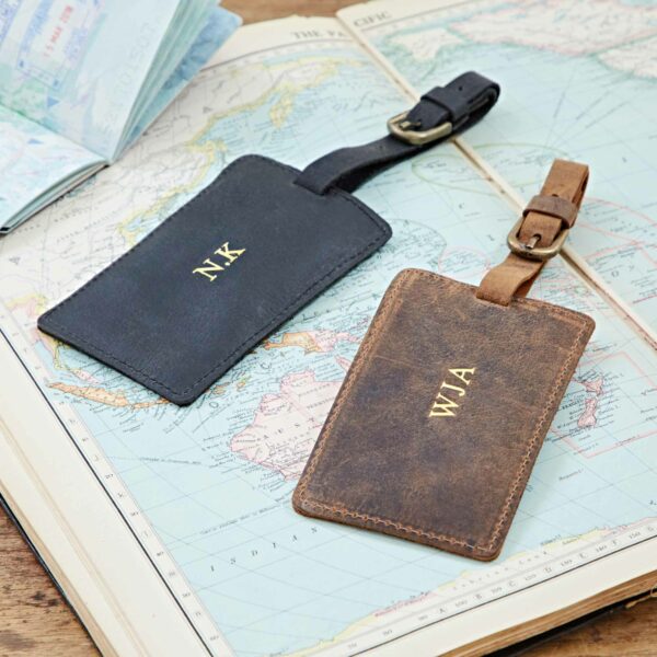 Two Personalised Buffalo Leather Luggage Tags, one black monogrammed “NK” and one brown with “WJA,” are displayed on an open atlas, with a passport partly visible in the background.
