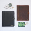Two Personalised Distressed Buffalo Leather Document Portfolios—one black with “JASMINE” and one brown with “T.P.”—are shown on a white surface with office supplies and a plant, highlighting their premium timeless style.