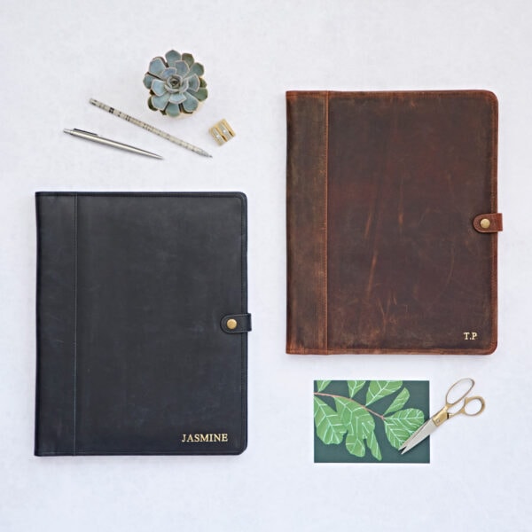 Two Personalised Distressed Buffalo Leather Document Portfolios—one black with “JASMINE” and one brown with “T.P.”—are shown on a white surface with office supplies and a plant, highlighting their premium timeless style.