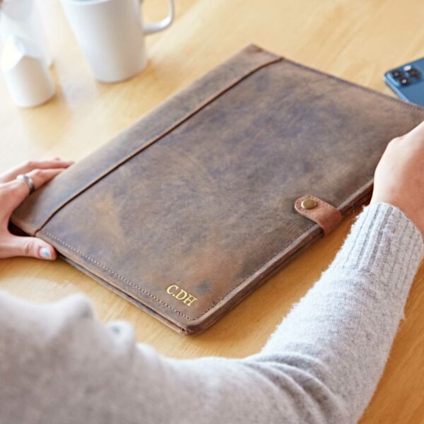 Person holding a personalised leather portfolio crafted from distressed buffalo leather with initials "CDF" embossed in gold, placed on a wooden table beside mugs and a mobile phone.