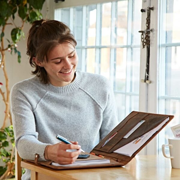 A person in a light sweater sits by a window, smiling as they write in a notebook and review papers stored in a Personalised Distressed Buffalo Leather Document Portfolio.