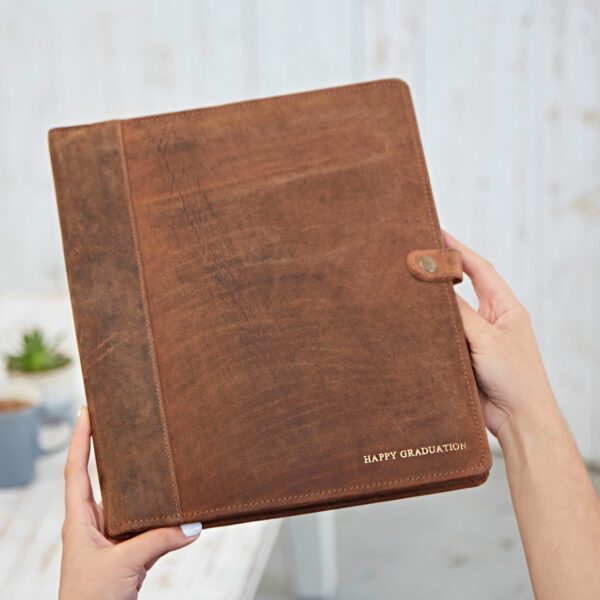 A person holds a Personalised Distressed Buffalo Leather Document Portfolio with a button closure and "HAPPY GRADUATION" embossed in gold on the bottom right corner.