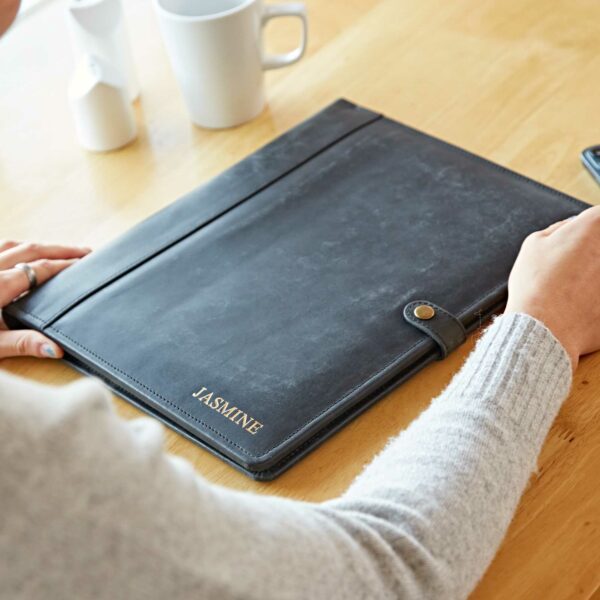 A person sits at a table with a Personalised Distressed Buffalo Leather Document Portfolio labeled "JASMINE," alongside a white mug, small containers, and a smartphone.