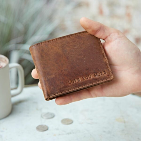 A hand holds a Personalised Buffalo Leather Billfold Wallet embossed with "LOVE YOU MORE," near a white mug and coins on a light surface.