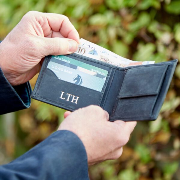 A person holds an open Buffalo Leather Wallet containing banknotes and cards labelled Lloyds Bank, John Lewis, and Waitrose, with the initials "LTH" on the personalised wallet.