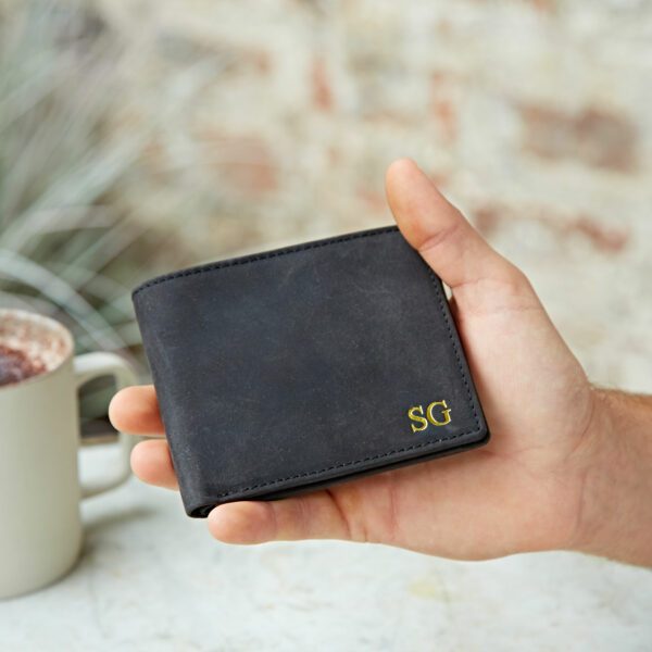 A hand holds a closed Personalised Buffalo Leather Billfold Wallet with gold "SG" initials embossed in the corner; a white mug is visible in the background.