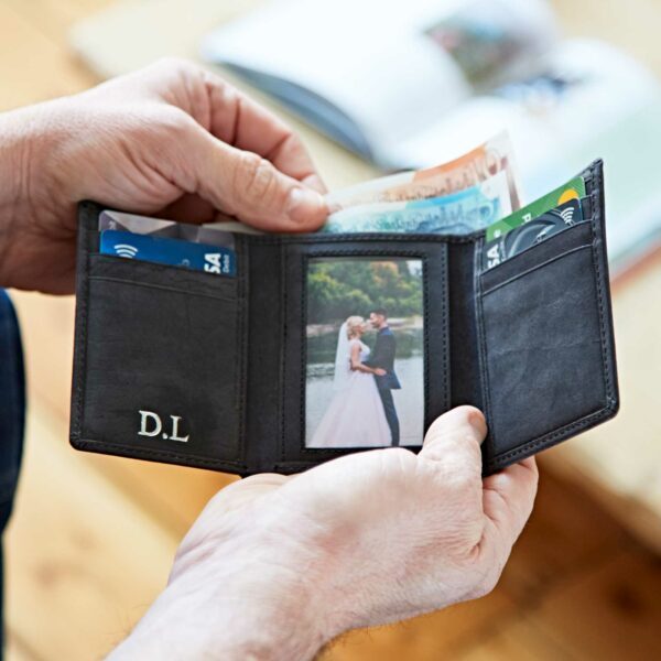 A person holds an open personalised wallet containing cash, cards, and a photo of a couple dressed in wedding attire by the water. The initials "D.L." are embossed on the wallet.