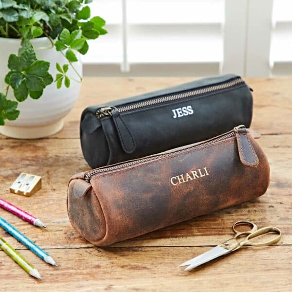 Two Personalised Buffalo Leather Round Pencil Cases, one black labeled "JESS" and one brown labeled "CHARLI," on a wooden desk with pencils, a sharpener, scissors, and a potted plant.