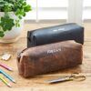 Two Personalised Buffalo Leather Square Pencil Cases—one brown labeled "JORDAN" and one black labeled "MRS PICKLES"—are on a wooden table with pencils, scissors, and a plant.
