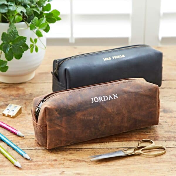 Two Personalised Buffalo Leather Square Pencil Cases—one brown labeled "JORDAN" and one black labeled "MRS PICKLES"—are on a wooden table with pencils, scissors, and a plant.