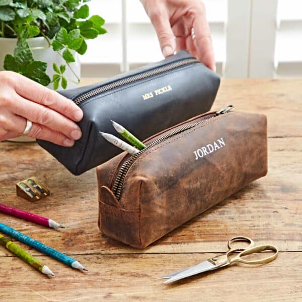 Two hands hold Personalised Buffalo Leather Square Pencil Cases—one black, one brown—reading "MR PICKLE" and "JORDAN," on a wooden table with stationery. The square design suits any desk or bag.
