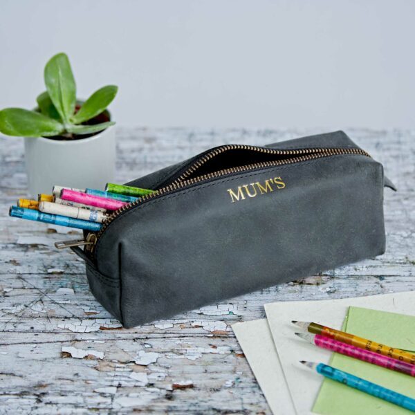 A grey personalised pencil case labelled "MUM'S" sits on a distressed wooden surface with coloured pencils inside, next to a small plant and some papers with pencils on them.