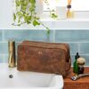 A Personalised Large Brown Buffalo Leather Wash Bag with the initials "R.L." rests on a bathroom counter beside a faucet, shaving brush, razor, small bottles, and a plant above.