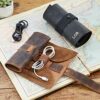 A Personalised Buffalo Leather Cable Organiser, filled with cables, sits open on a map beside a rolled-up version embossed with "LOR," while a loose cable and small plant complete this travel-ready setup.