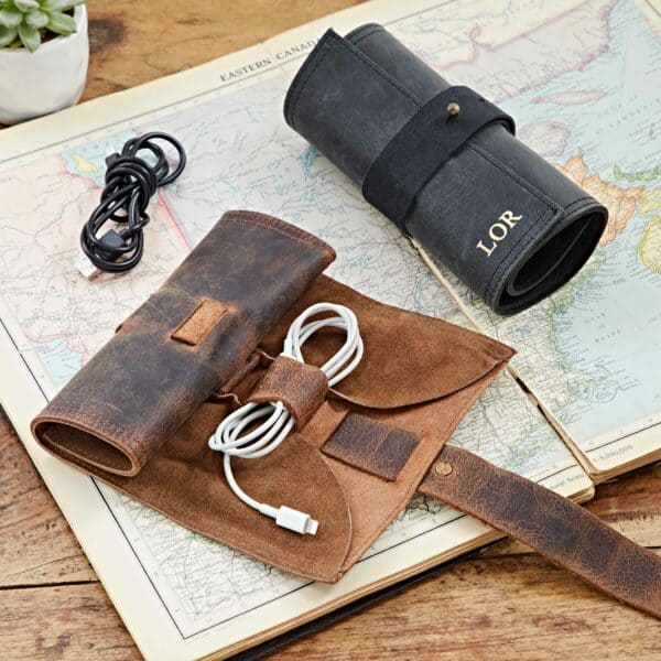 A Personalised Buffalo Leather Cable Organiser, filled with cables, sits open on a map beside a rolled-up version embossed with "LOR," while a loose cable and small plant complete this travel-ready setup.