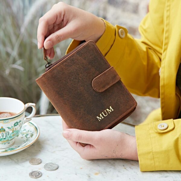 A person in a yellow coat holds a Personalised Brown Buffalo Leather Purse labeled "MUM" beside a table with a teacup, saucer, and coins.
