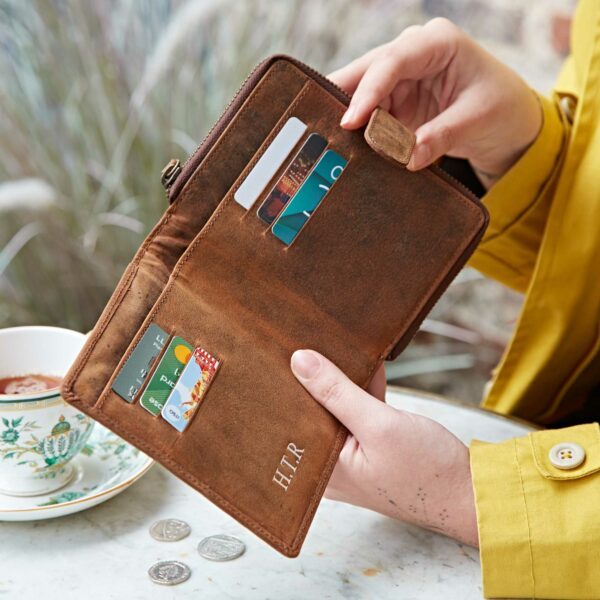 A person in a yellow coat holds an open Personalised Brown Buffalo Leather Purse displaying multiple cards near a teacup and some coins on a marble table.