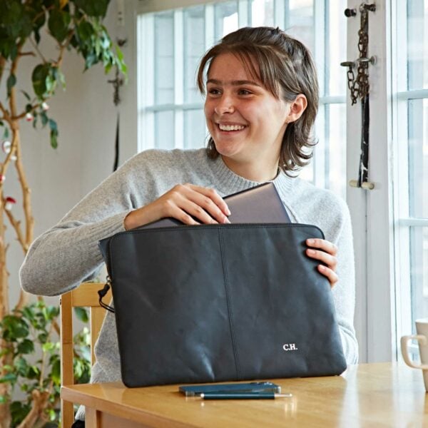 A person sits at a table and smiles while placing a tablet into a black zippered Personalised Buffalo Leather 14 inch Laptop Case. Nearby are a smartphone and a pen, with windows and a potted plant visible in the background.
