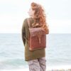 A person with long curly hair in a green sweater stands near water, facing away, wearing a Personalised Brown Buffalo Leather Slim Backpack.