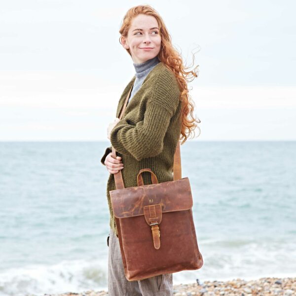A woman with long red hair stands on a pebbled beach, wearing a green cardigan and gray turtleneck, holding a Personalised Brown Buffalo Leather Slim Backpack.