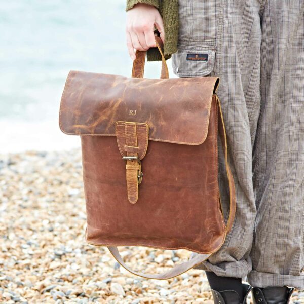 Someone stands on a rocky beach holding the Personalised Brown Buffalo Leather Slim Backpack, featuring a slim design, buckle closure, and the initials "RJ" on the flap.