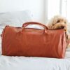 A curly-haired dog rests its head on a Personalised Brown Leather Duffle Style Gym Bag, which sits on a bed.