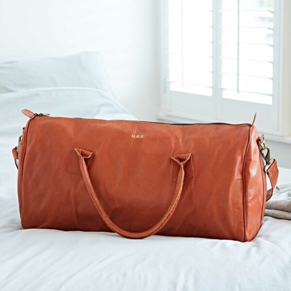 A Personalised Brown Leather Duffle Style Gym Bag with handles sits on a white bed near a window with white shutters.