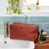 A Personalised Large Brown Leather Wash Bag sits on a bathroom sink beside a razor, shaving brush, and toiletries, with green tiles and a plant in the background.