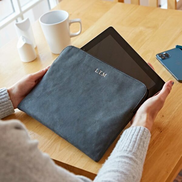 Someone puts a tablet into a Personalised Buffalo Leather Tablet Case on a wooden table, with a cup, two containers, and a smartphone nearby.